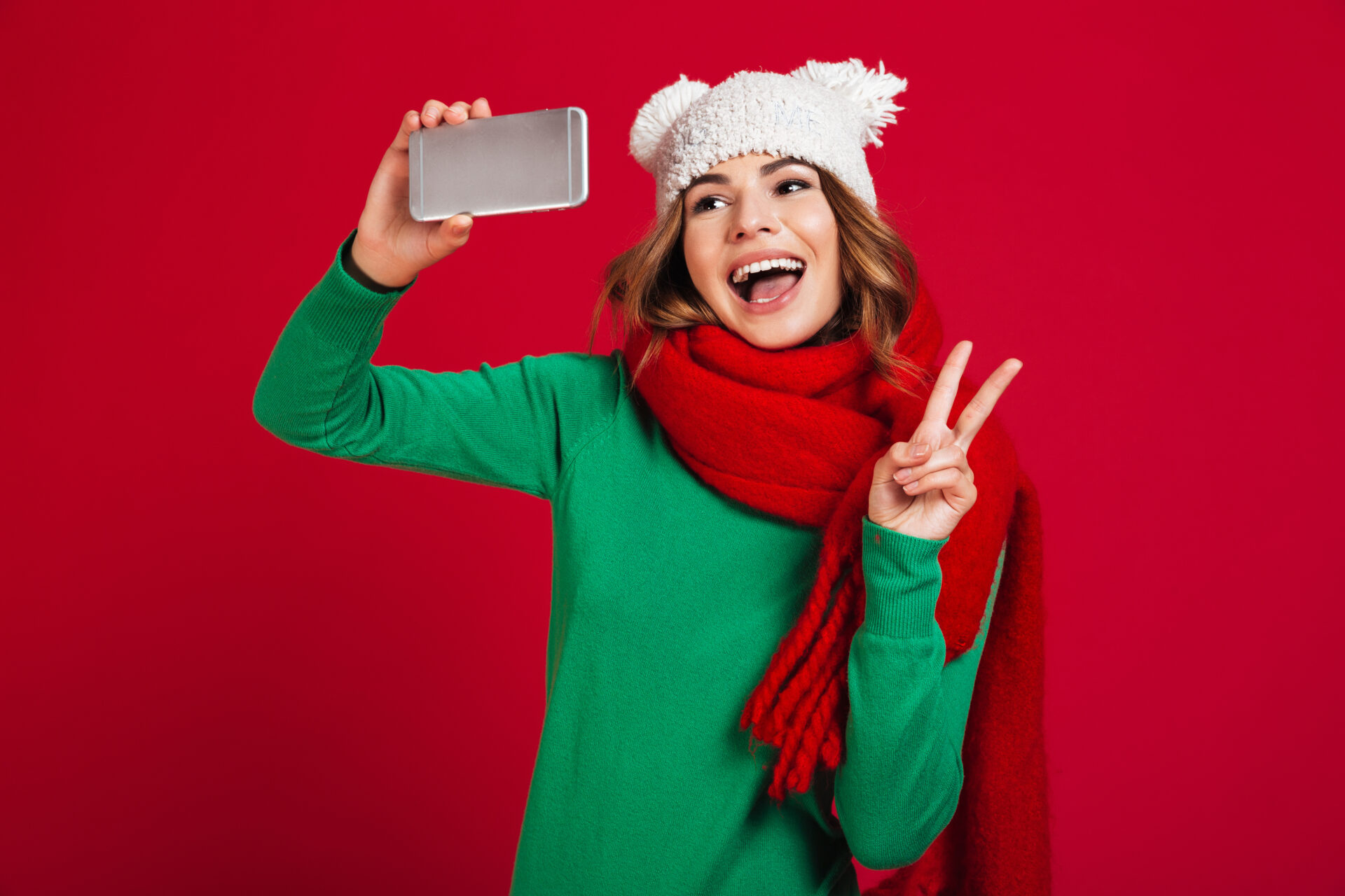 Photo of happy young woman wearing hat and scarf. Looking aside make selfie by phone showing peace gesture.